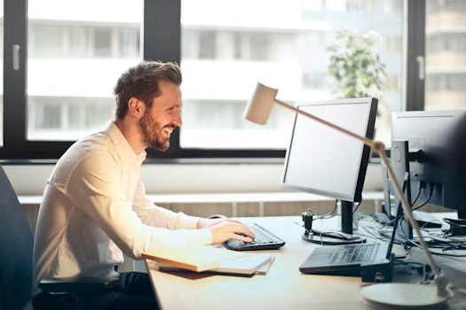 pexels photo 840996 840996 A man smiling while working at an office desk with a computer and natural daylight streaming in through large windows.